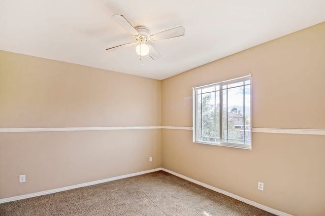 Empty carpeted bedroom with tan walls, a white chair rail, a ceiling fan and a window looking out to trees and neighboring rooftops