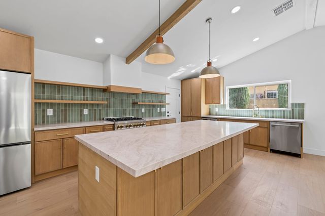 Claremont kitchen with oak cabinets and open shelves, a green vertical tile backsplash, a long stone-topped island, two hammered metal pendants and a wood beam across a vaulted ceiling.