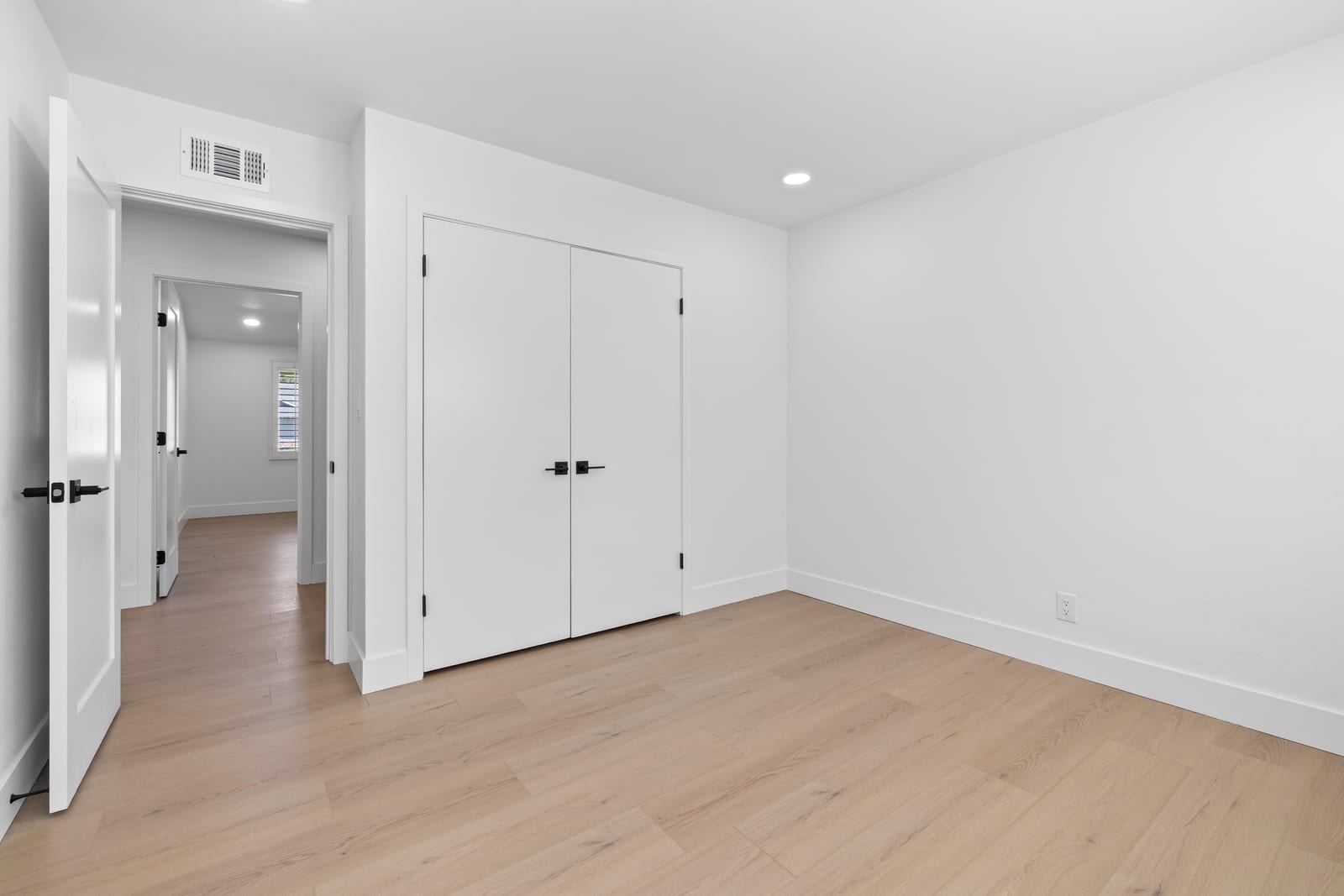 Empty bedroom in a Claremont house with white walls, light wood plank floors, white double closet doors with black handles and an open door to a hallway.