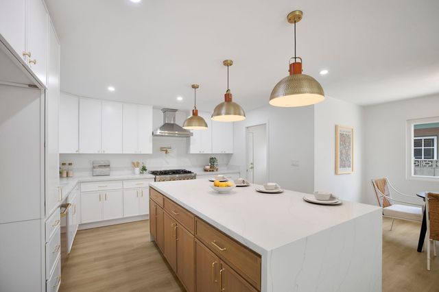 Remodeled kitchen with white shaker cabinets, a wood island with a white waterfall stone top, three hammered brass pendant lights and a range under a steel hood.