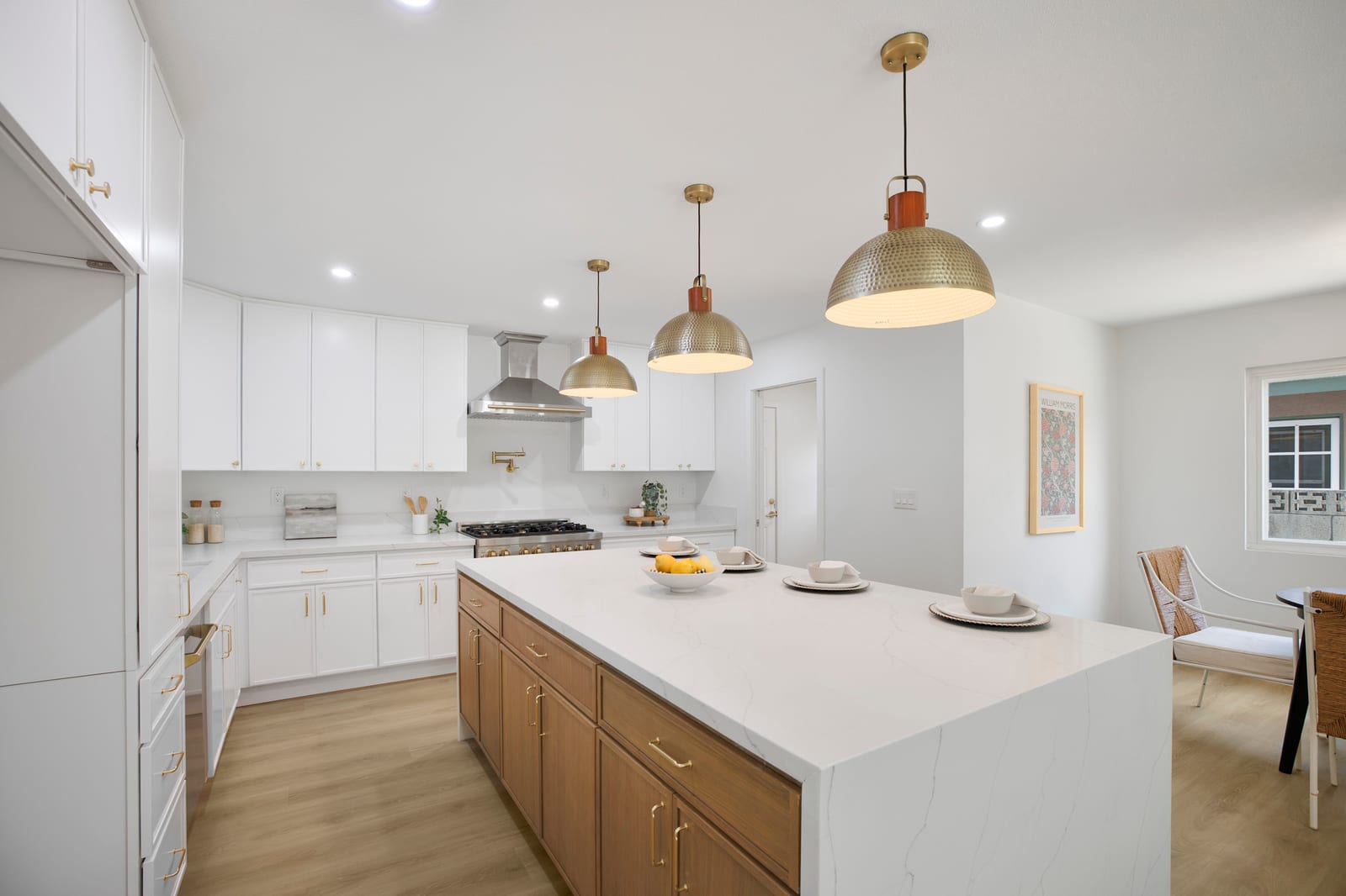 Remodeled kitchen with white shaker cabinets, a wood island with a white waterfall stone top, three hammered brass pendant lights and a range under a steel hood.