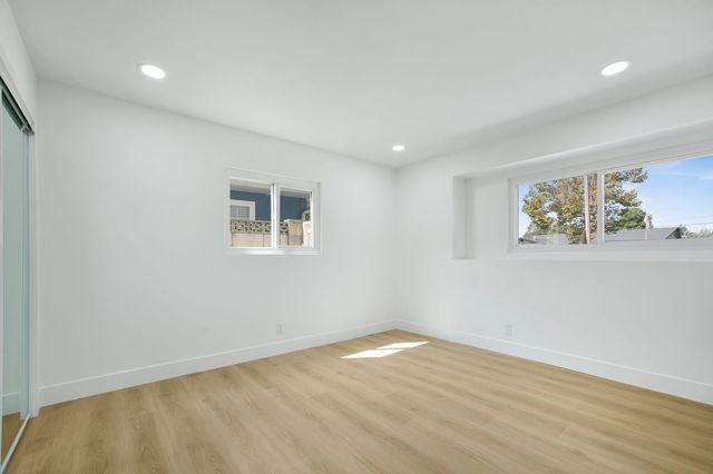 An empty bedroom with white walls, recessed ceiling lights, light wood-look flooring, a mirrored closet door and two sliding windows letting in daylight