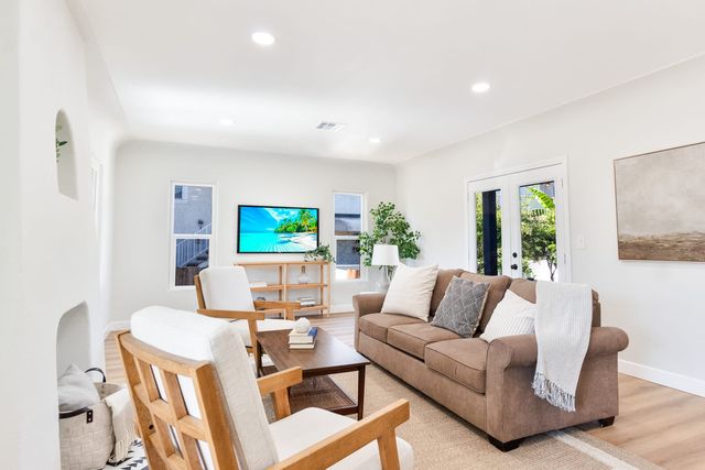 A bright living room with a brown sofa, two wood-framed armchairs, a coffee table on a woven rug, a wall-mounted television and French doors to a leafy patio