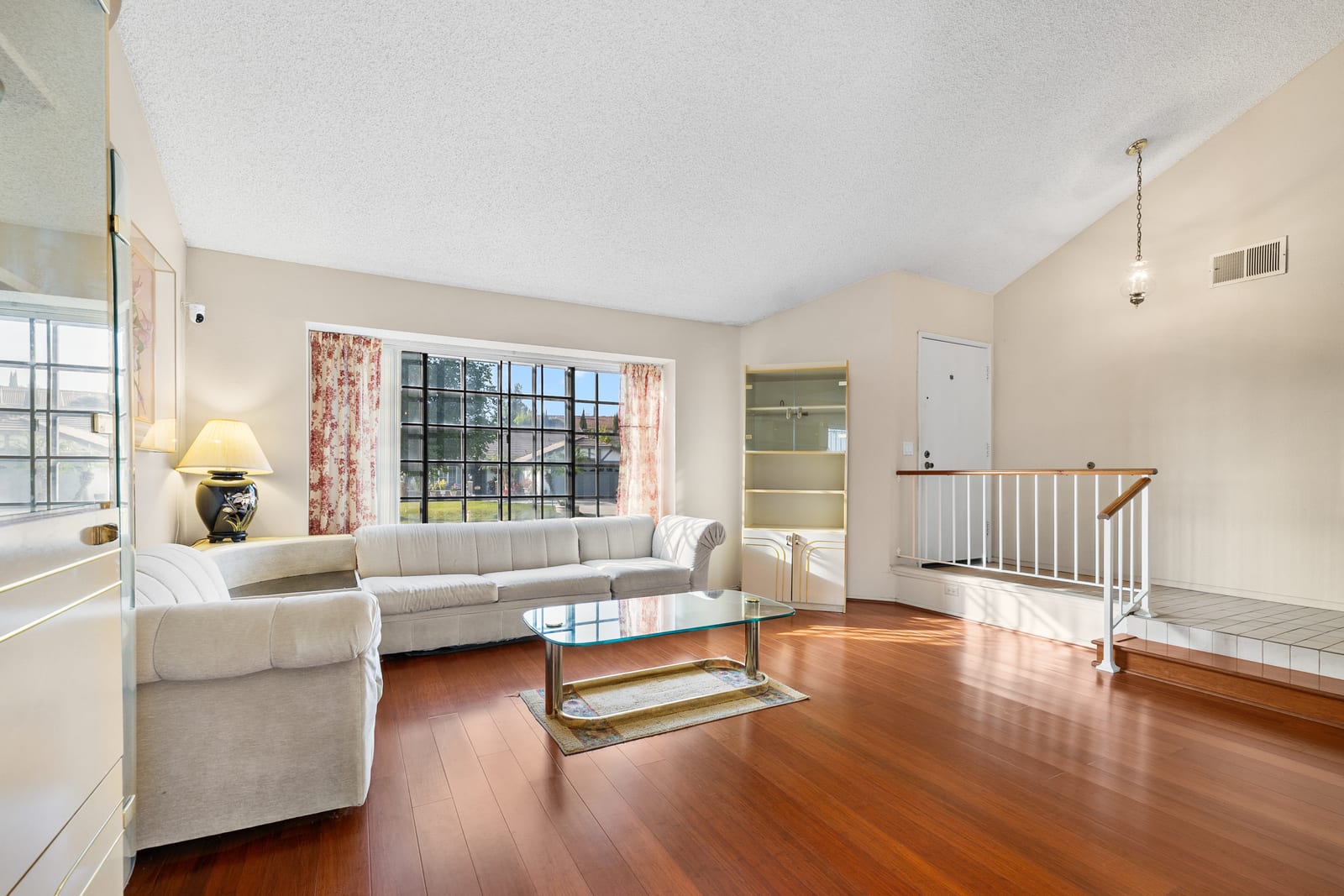 A living room with a vaulted textured ceiling, a cream sectional sofa, a glass coffee table, cherry-toned wood floors and a large gridded window, with a few steps up to the entry