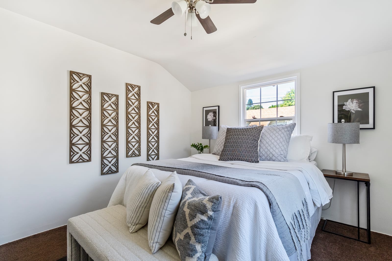 A staged bedroom in an Ontario house with a sloped white ceiling, a ceiling fan, a made bed with gray and white pillows, two table lamps and a single window.