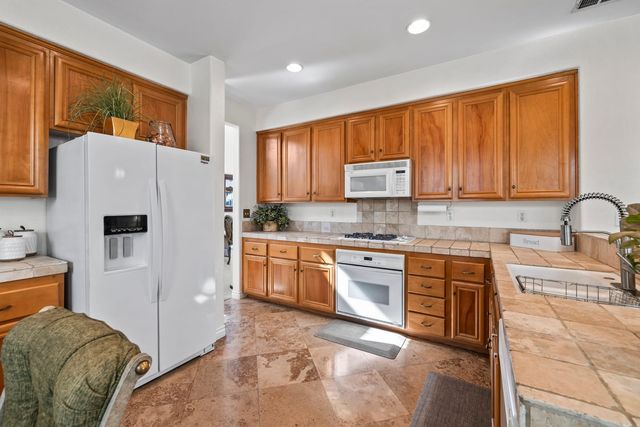 A kitchen in a Corona house with honey-toned wood cabinets, tiled counters and backsplash, a white side-by-side refrigerator, a white wall oven and microwave, and a travertine floor.