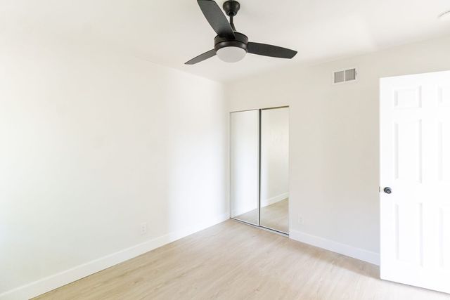 An empty bedroom in a West Covina house with white walls, light wood-look flooring, a dark three-blade ceiling fan, a mirrored sliding closet and an open white panel door.