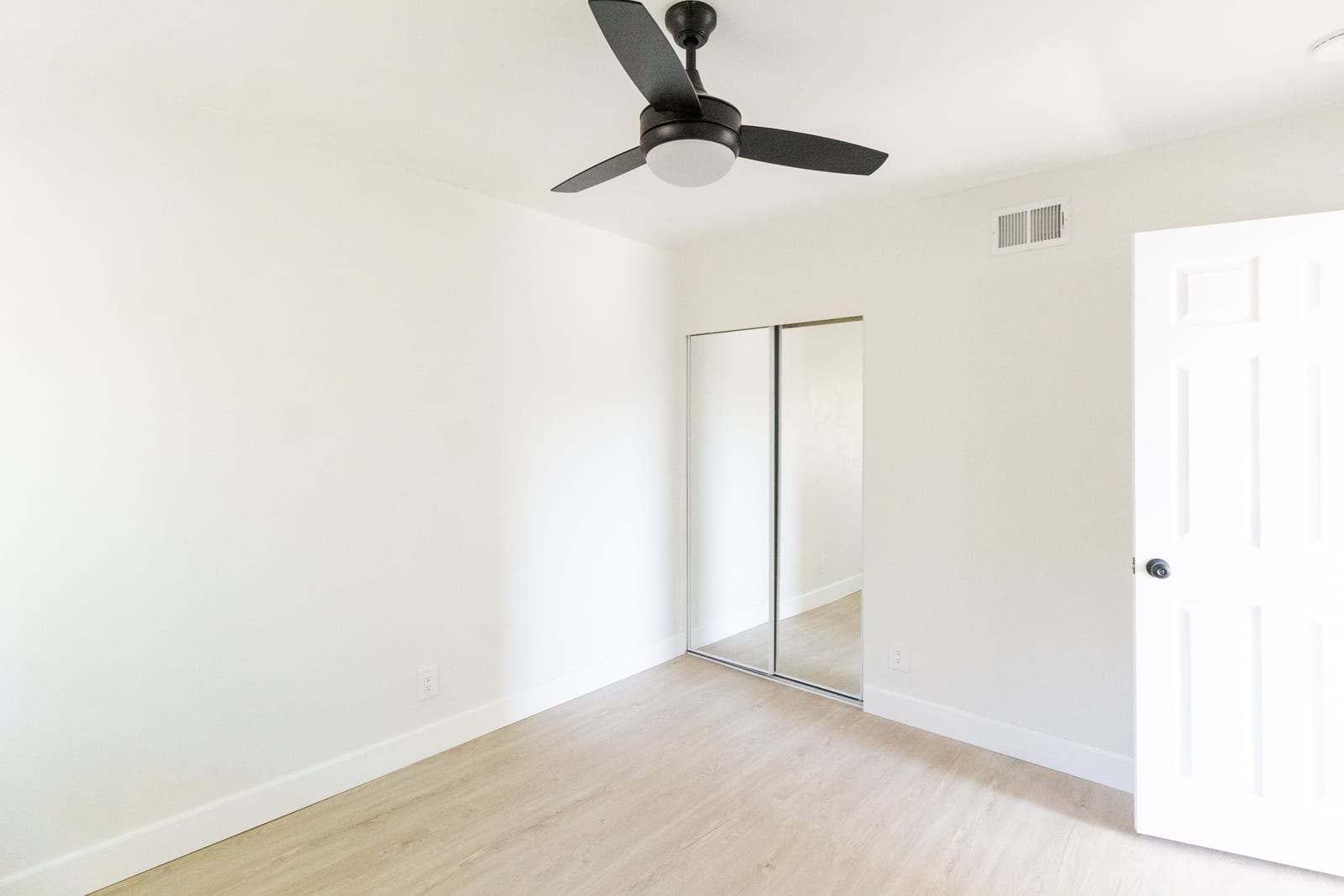 An empty bedroom in a West Covina house with white walls, light wood-look flooring, a dark three-blade ceiling fan, a mirrored sliding closet and an open white panel door.