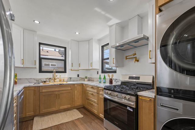A renovated U-shaped kitchen with white upper cabinets, wood lower cabinets, gold hardware, marble-look counters, a gas range and a stacked washer and dryer.