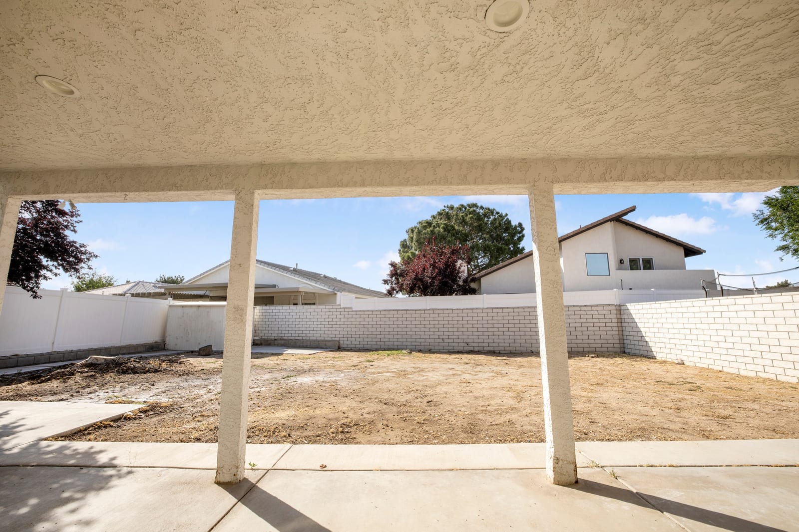 A covered concrete patio with stucco columns looking out on a bare dirt backyard enclosed by block and vinyl fencing under a blue sky.