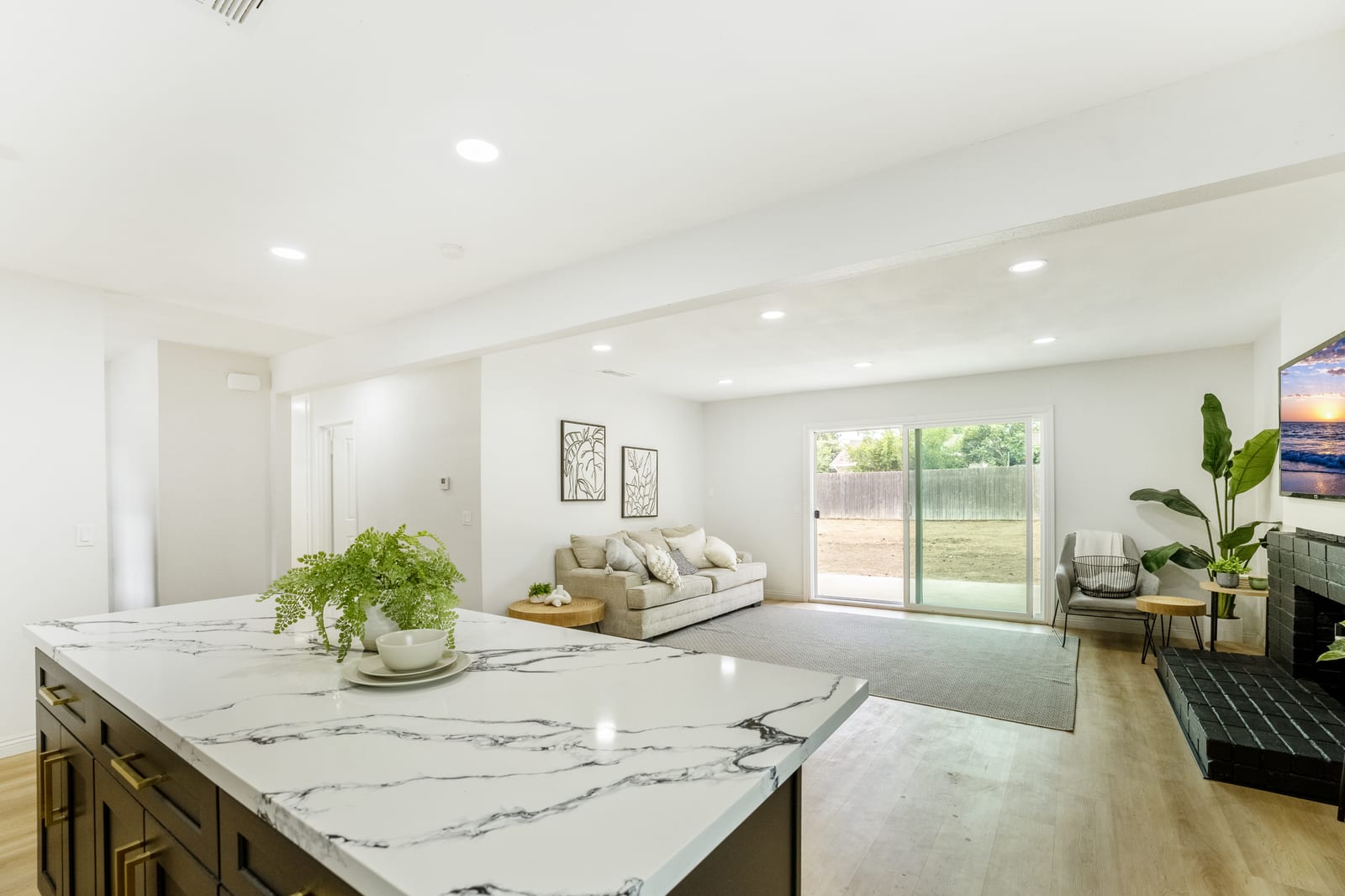 An open living area seen from a marble-look kitchen island, with a sofa, a fireplace painted black and a sliding glass door to the backyard.