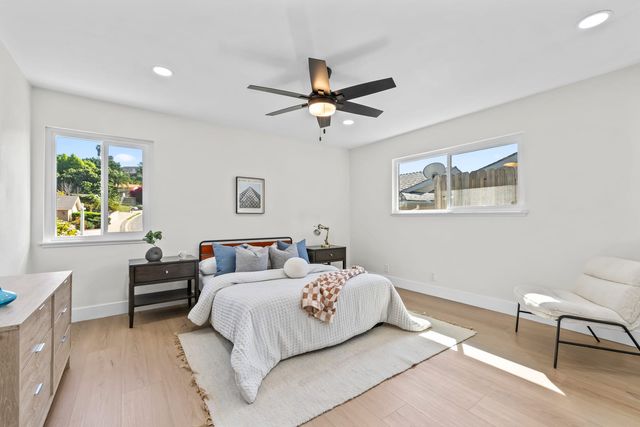 A staged bedroom in a Diamond Bar house with light wood floors, a ceiling fan, a made bed and two windows looking out to a hillside street and a backyard fence.