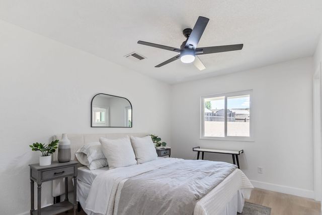 A staged bedroom in a Rialto house with a made bed, gray nightstands, an arched mirror, a ceiling fan and a window looking out to a block wall.