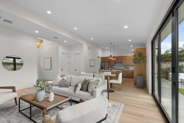 An open living, dining and kitchen space in a Costa Mesa house, with wood cabinets, light flooring and black-framed glass doors opening to a patio
