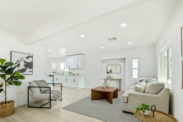 A renovated open living room and white kitchen in a San Bernardino house, with a ceiling air vent, recessed lights and light wood-look flooring