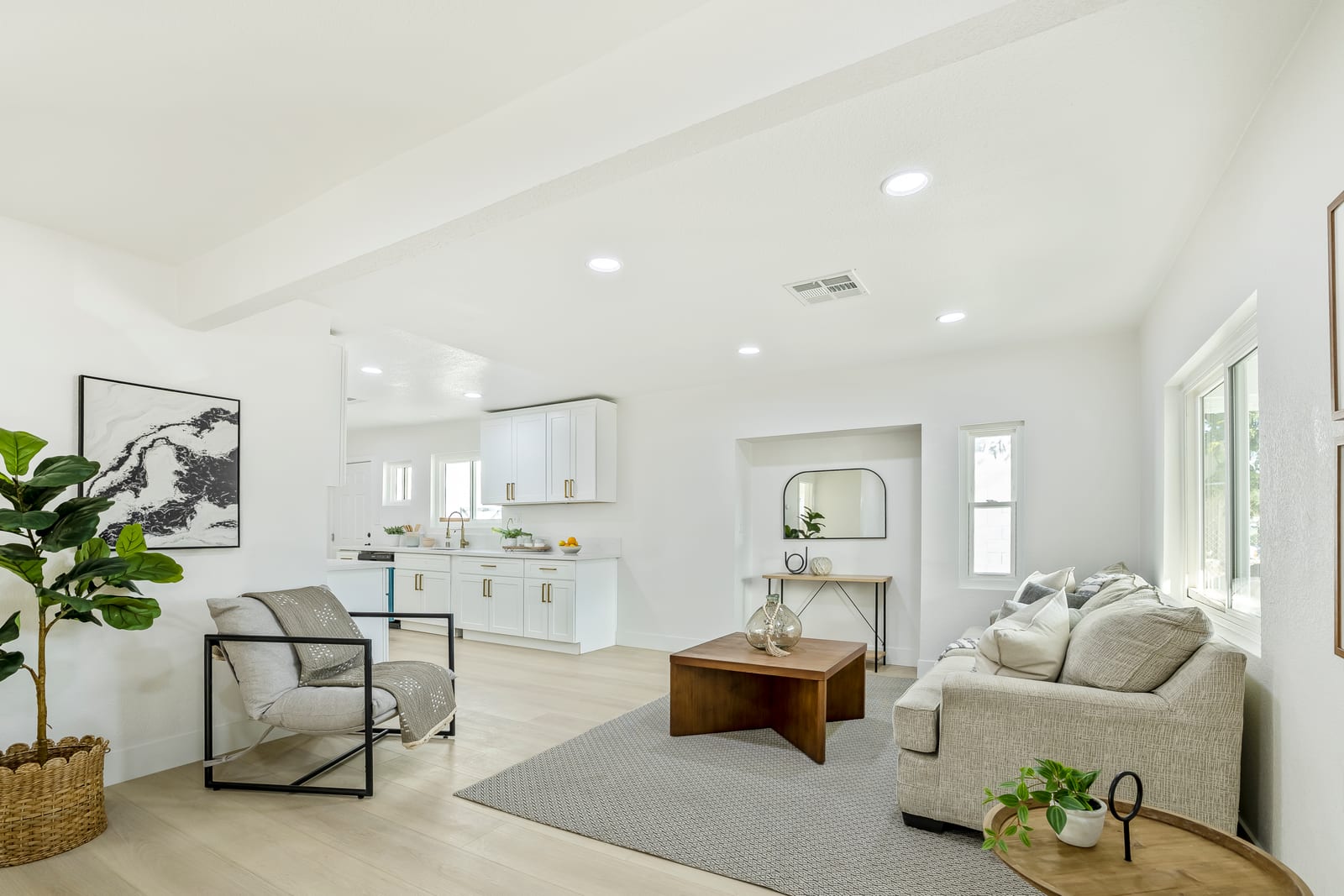 A renovated open living room and white kitchen in a San Bernardino house, with a ceiling air vent, recessed lights and light wood-look flooring