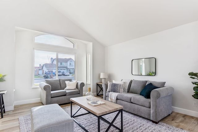 A staged living room in a Rialto house with a vaulted ceiling, two gray sofas, a wood-look floor and a front window looking out on a residential street