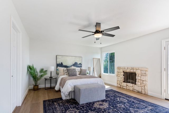 A staged Rancho Cucamonga bedroom with a made bed, a blue rug, a ceiling fan and a stacked-stone fireplace beside a window