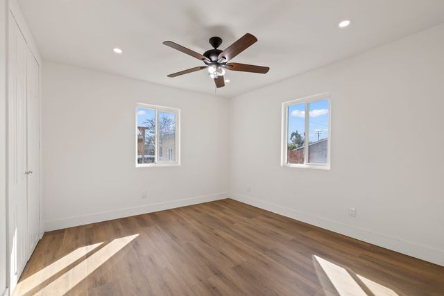 An empty bedroom with white walls, wood-look plank flooring, a five-blade ceiling fan, closet doors on the left and two windows letting in afternoon sun