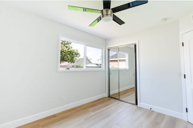 An empty bedroom with light gray walls, pale wood-look flooring, a black-bladed ceiling fan, a mirrored sliding closet and a window looking out at a tree and neighboring rooftops