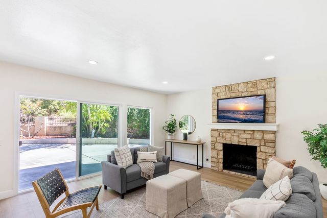 A staged living room with gray sofas, woven ottomans, a stone fireplace with a wall-mounted screen, and sliding glass doors opening to a patio and garden