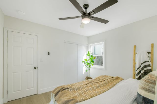 A small bedroom with white walls, a five-blade ceiling fan, a paneled door, a sliding closet and a potted plant beside a sunny window, with a striped throw on the bed