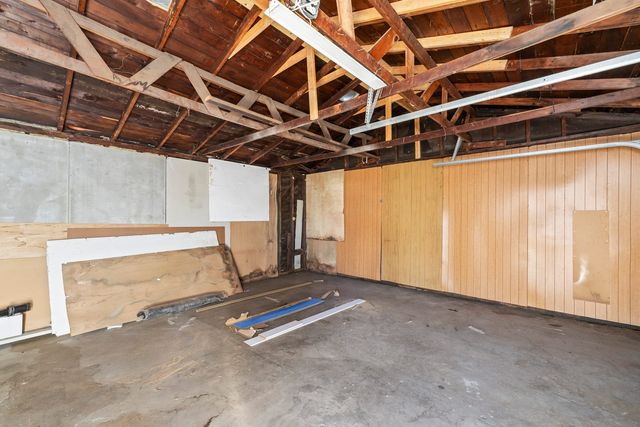 An unfinished garage interior with exposed roof framing, mixed wall paneling and leftover boards leaning on a bare concrete floor