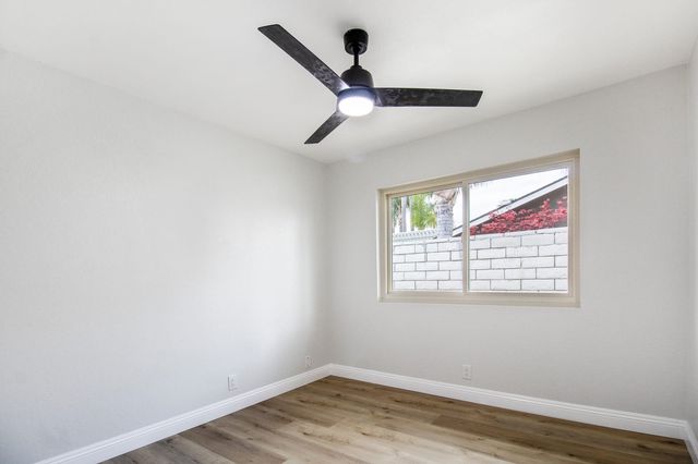 An empty bedroom with pale gray walls, wood-look flooring, a three-blade ceiling fan and a sliding window that looks out on a block wall and red foliage
