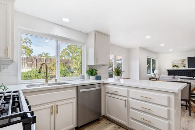 Bright kitchen with white cabinets, brass pulls, quartz counters, a stainless dishwasher and a window over the sink facing a fenced backyard