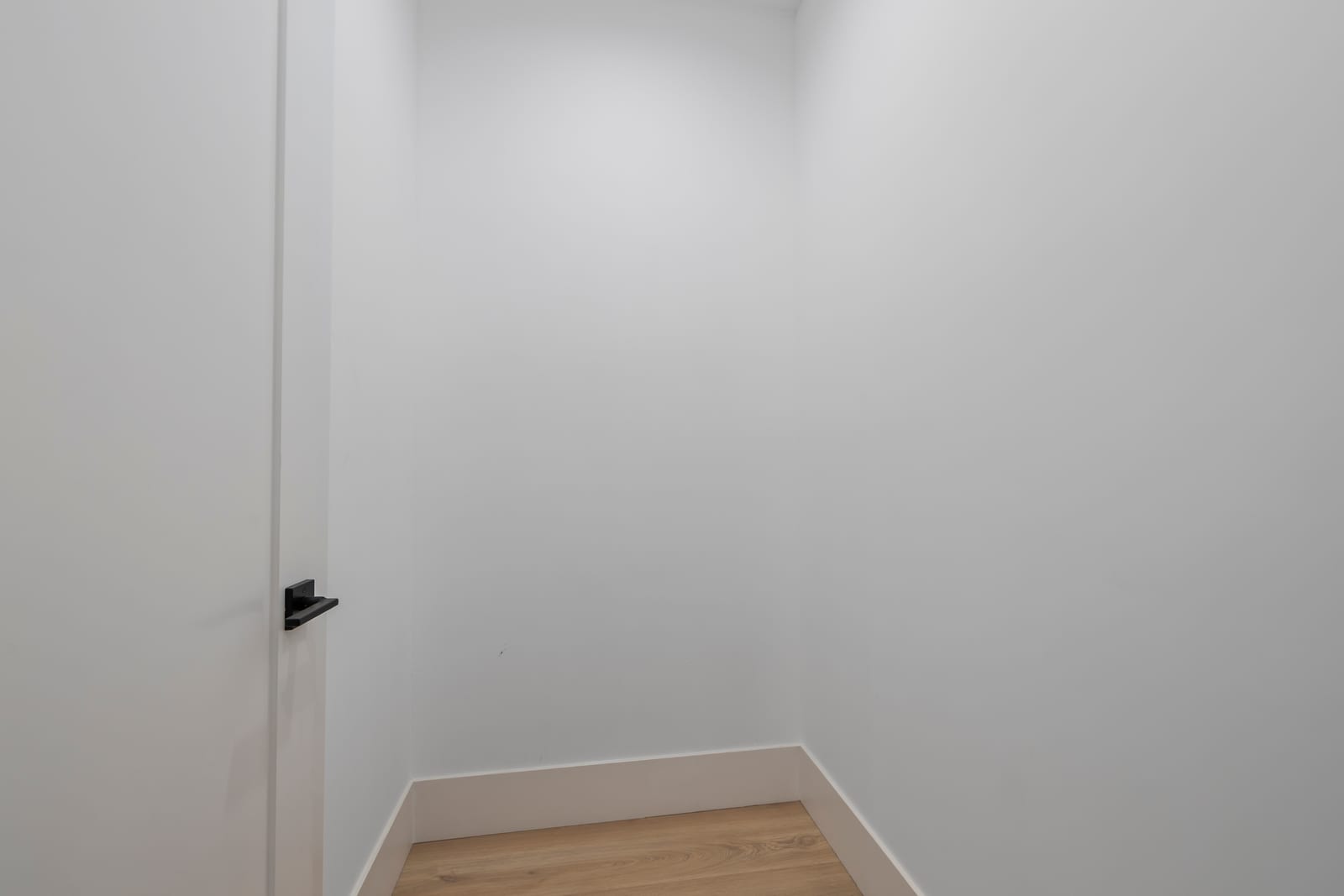 Small empty room with white walls, an open white door with a black lever handle and light wood flooring