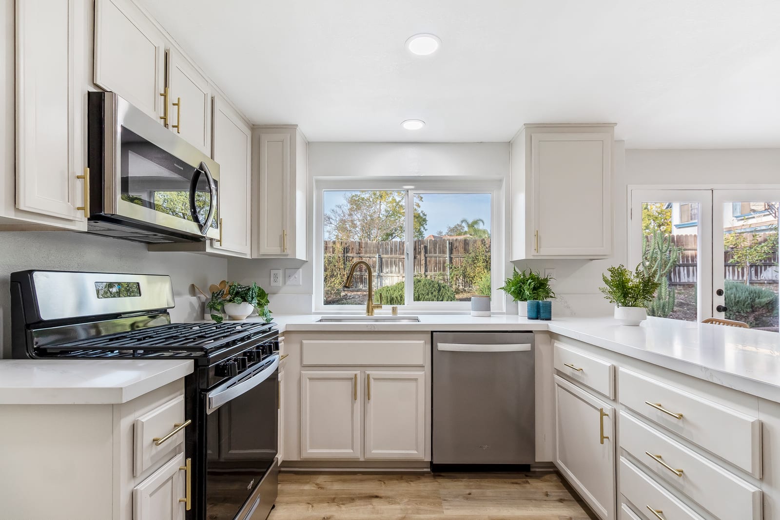 White kitchen with brass cabinet pulls, a black and stainless gas range, a window over the sink facing a fenced backyard, and potted plants on the counters