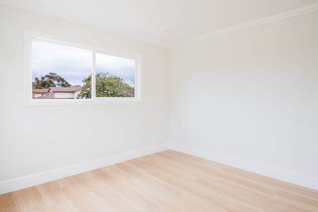 Empty bedroom with light wood plank flooring, white walls and a wide sliding window looking out at trees and neighboring rooftops