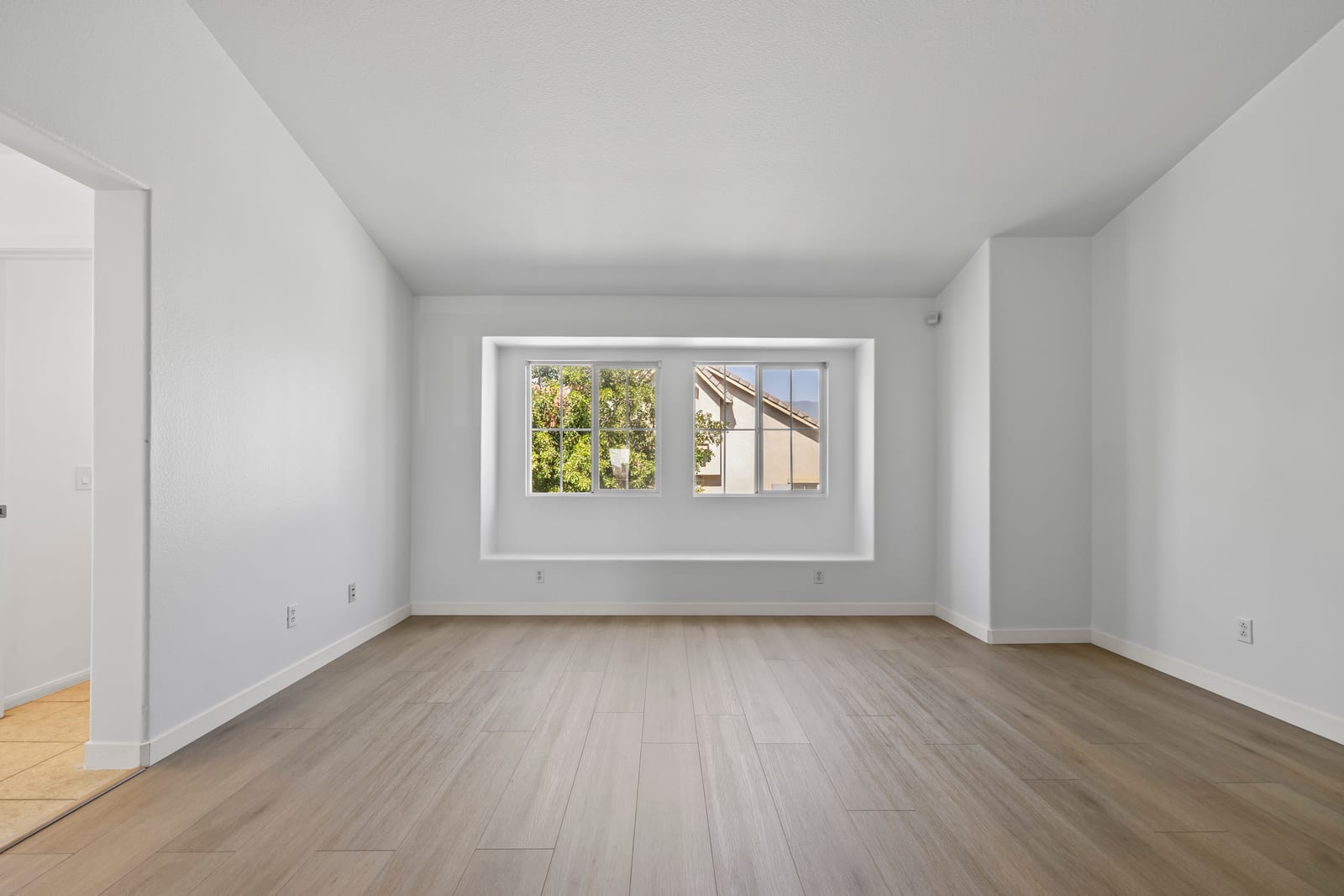 Empty living room with light wood-look flooring, white walls, a vaulted ceiling and a pair of windows in a deep recessed alcove