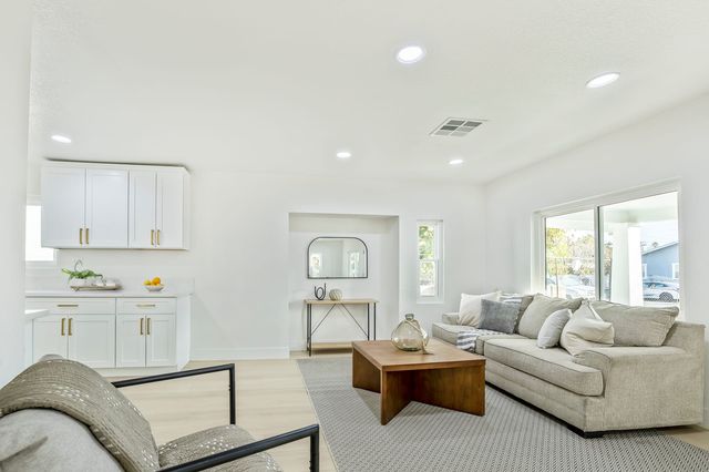 Bright staged living room with a beige sectional, a wood coffee table, white shaker kitchen cabinets with brass pulls and a sliding glass door to a covered porch