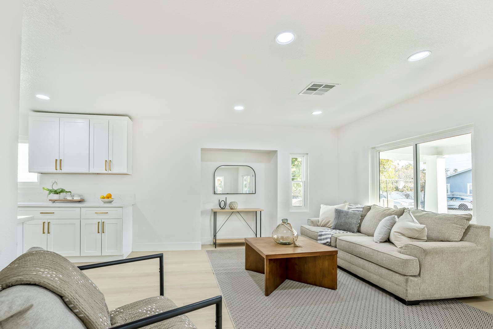 Bright staged living room with a beige sectional, a wood coffee table, white shaker kitchen cabinets with brass pulls and a sliding glass door to a covered porch