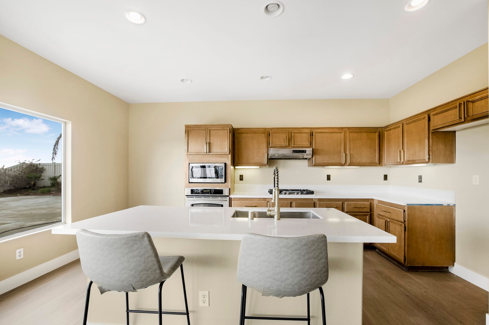 Vacant kitchen in a San Bernardino house with wood cabinets, a white quartz island with two gray stools and a large window onto a bare backyard.
