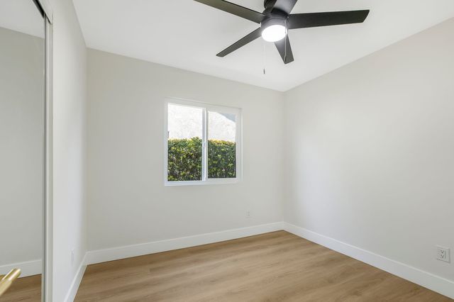 Empty bedroom in a Rancho Cucamonga house with new plank flooring, a black ceiling fan, a mirrored closet door and a window onto a green hedge.