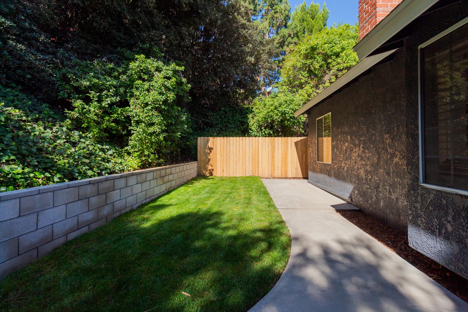 Side yard of a Rancho Palos Verdes house: a fresh lawn between a block retaining wall and a concrete path, a new wood fence and mature trees behind it.