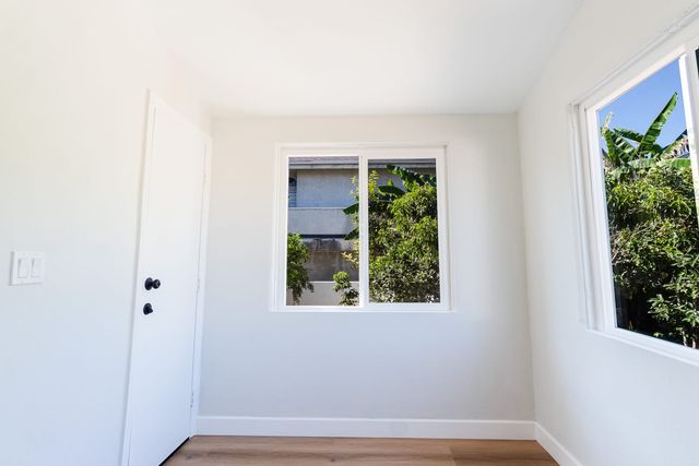 Empty, freshly painted bedroom in a Whittier house, with a white door, wood-look flooring and two sliding windows looking out on banana plants and a neighboring building.