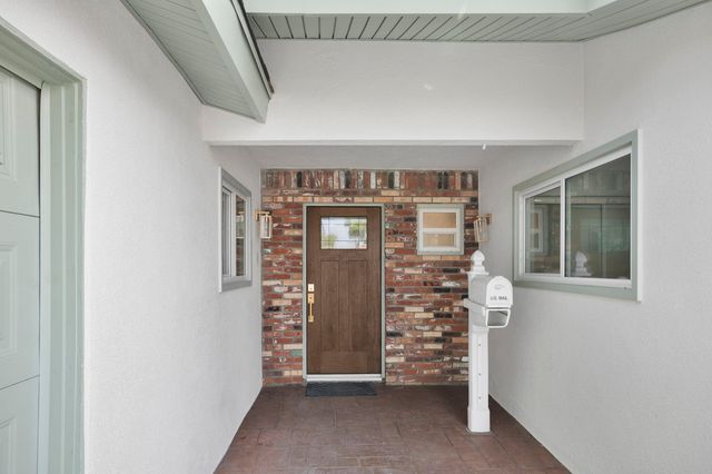 Covered front entry of a Santa Ana house: a wood front door set in a brick wall, brass lantern lights, a white mailbox post and sliding windows on stucco walls.