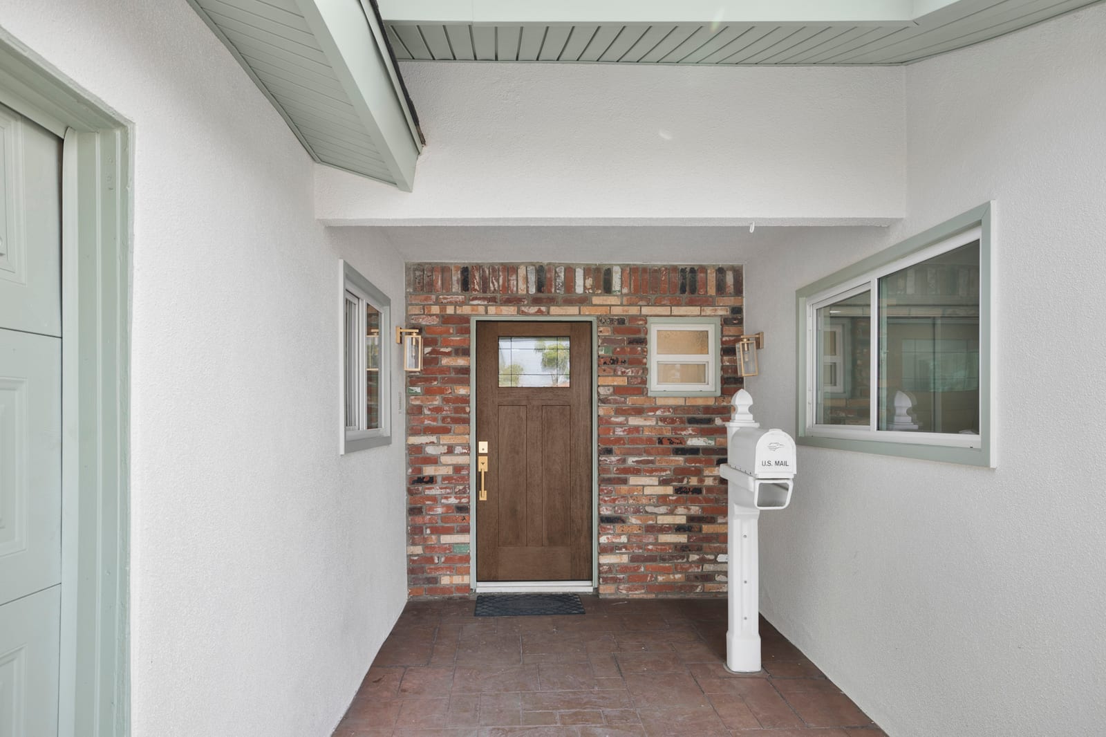 Covered front entry of a Santa Ana house: a wood front door set in a brick wall, brass lantern lights, a white mailbox post and sliding windows on stucco walls.