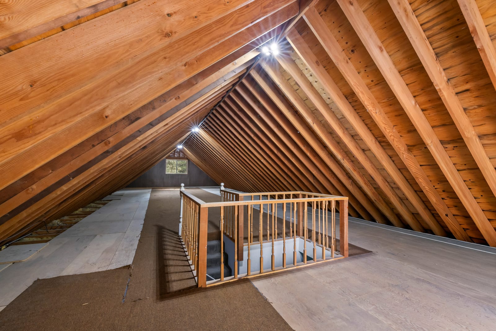 An open attic loft under a steep wood-plank roof with exposed rafters, a wooden railing around the stair opening and a small window at the gable end.