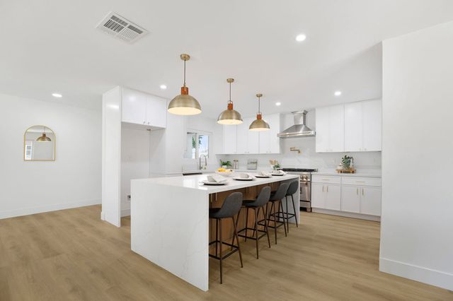 A renovated open kitchen with white shaker cabinets, a white waterfall island with four gray bar stools, brass dome pendant lights and light oak-look flooring.