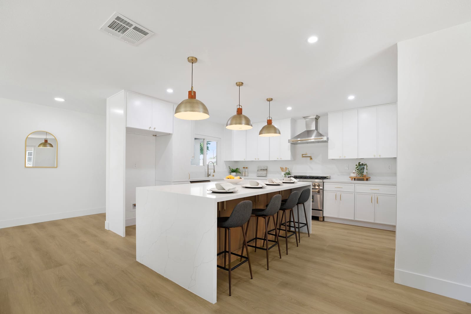 A renovated open kitchen with white shaker cabinets, a white waterfall island with four gray bar stools, brass dome pendant lights and light oak-look flooring.