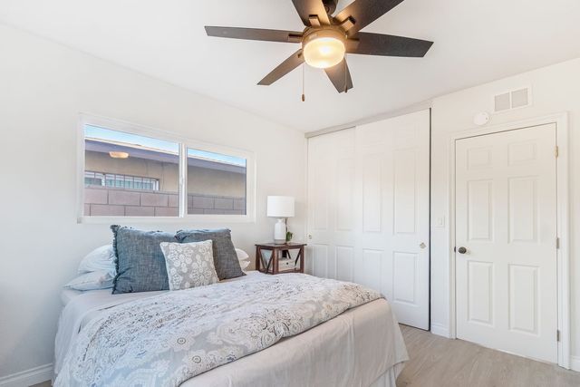 A bright staged bedroom with a made-up bed, gray and lace pillows, a dark ceiling fan, a nightstand lamp, white sliding closet doors and a wide window.