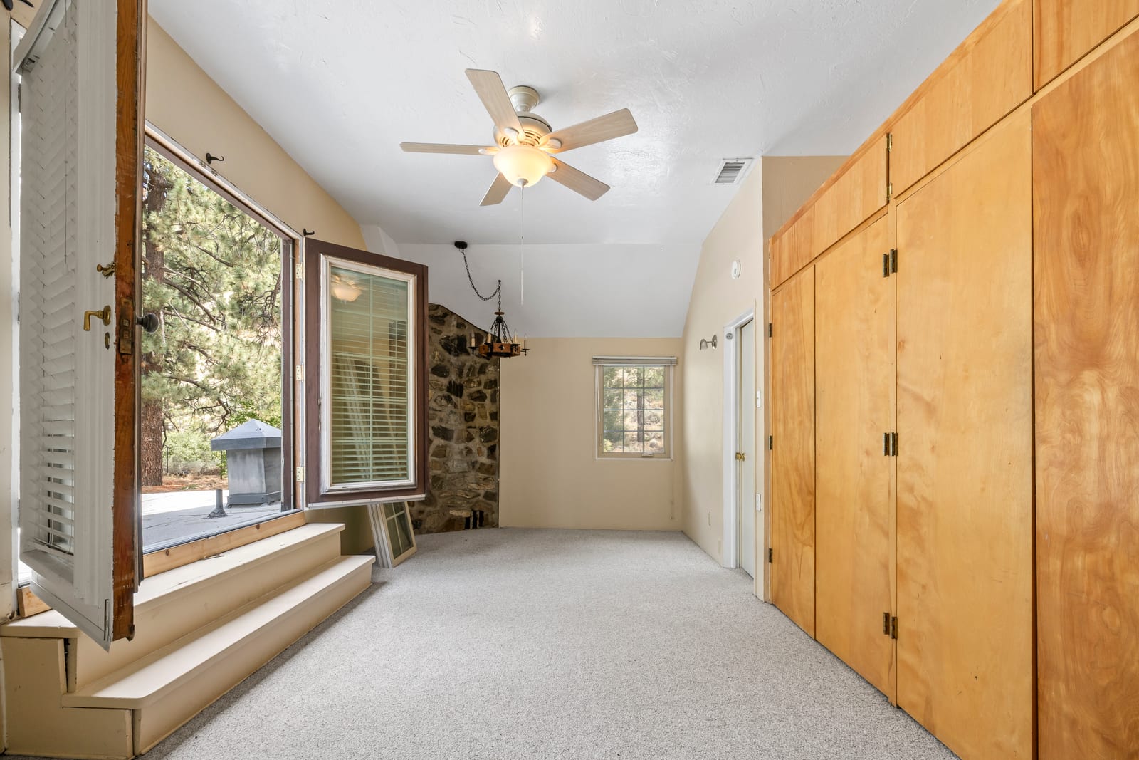 An empty carpeted room with a sloped ceiling, a ceiling fan, a wall of built-in wood cabinets, a stone wall in the far corner and tall open windows looking out to pine trees.