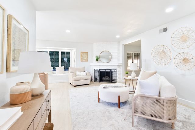 A bright staged living room with a white painted brick corner fireplace, a round mirror, a cane-sided sofa, a round ottoman, light wood floors and shuttered windows.