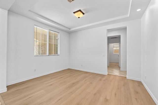 An empty bedroom with white walls, a tray ceiling with a flush light, wood-look plank floors and a window with blinds, with a doorway leading to a tiled hallway.