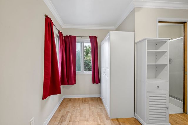 An empty bedroom with red curtains on two windows, light wood floors, a tall white wardrobe and a white open shelving cabinet, with a doorway to a bathroom with a glass shower.