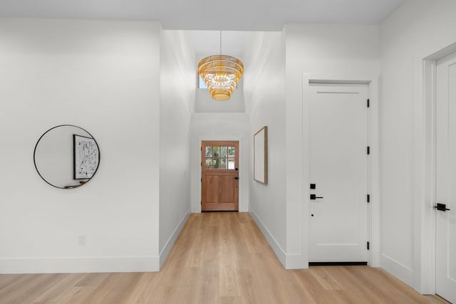 Bright entry hallway in a Costa Mesa home with white walls, light wood-look plank flooring, a tiered gold chandelier under a high ceiling and a wood front door with divided-light glass